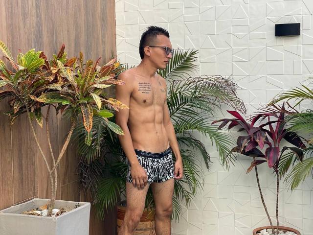 Shirtless WorldSexxxADS with tattoos, wearing black and white zebra-patterned briefs, standing next to potted plants, wearing glasses, has a slim, toned build, and short dark hair.