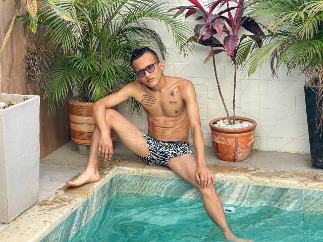 WorldSexxxADS with a fit, tan, muscular, tattooed body, wearing black and white patterned swim trunks, sitting by a small pool, legs spread, one foot in water, smiling.
