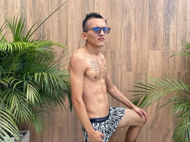WorldSexxxADS with a slim, shirtless build, wearing zebra-patterned shorts and blue sunglasses, standing with one leg raised, tattoos on chest, short black hair, and green potted plants.