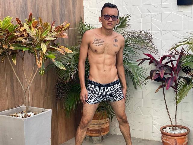 WorldSexxxADS, wearing black and white striped speedos, standing with a muscular, tan, and hairy chest, tattoos on right pectoral, and dark sunglasses, surrounded by potted plants.