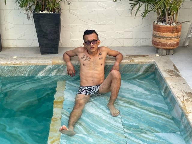 WorldSexxxADS is a slim, tattooed dude with dark hair, wearing black-and-white striped swim trunks, sunglasses, and sitting with one leg up in a marble-tiled pool.