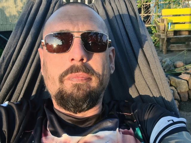 Martinleck54-hot with bald head, beard, sunglasses, and black shirt, lying back with dark cloth draped over head, sunlight on face, wearing a black and white sports shirt.