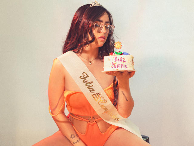Eva McAllister mit langen roten Haaren, Brille, Tiara, orangefarbenem Einteiler, weißem Gürtel, hält eine Torte mit der Aufschrift "Feliz Cumpleaños" und bunten Kerzen, sitzt, hat ein Tattoo am rechten Unterarm..