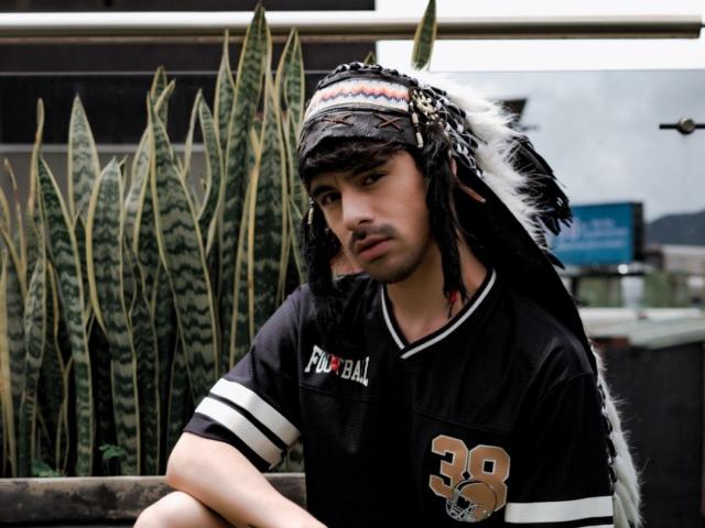 BryCamX with black sports jersey, number 38, wearing a feathered headband, looking slightly bored, sitting in front of tall green and yellow snake plants.