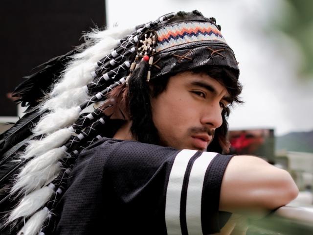 BryCamX with black and white feathered headpiece, dark hair, and black shirt with white stripes, looking to the side with a serious expression.