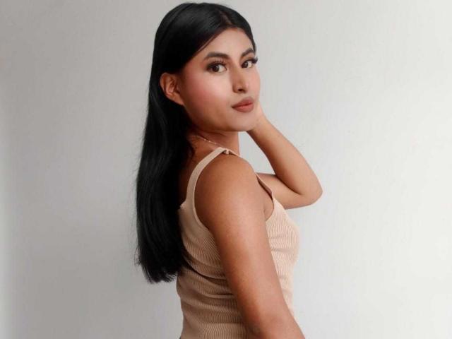 MichelleDenise with long black hair, brown eyes, and a tan ribbed tank top, looking over her shoulder, hand on neck, cute, small ass, subtle makeup, neutral expression.