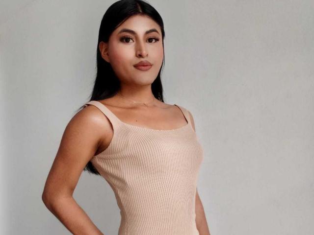 MichelleDenise has long black hair, tan skin, and is wearing a ribbed, beige tank top. Her small, perky tits are visible. She's standing with a neutral expression.