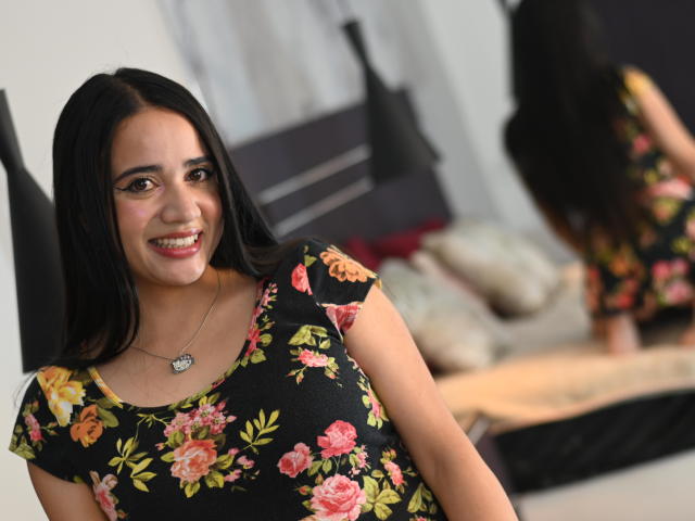 NicoleHolmes with long black hair, smiling, wearing a black floral dress, heart necklace, and visible tits in a mirror reflection. Her ass is slightly visible in the background.