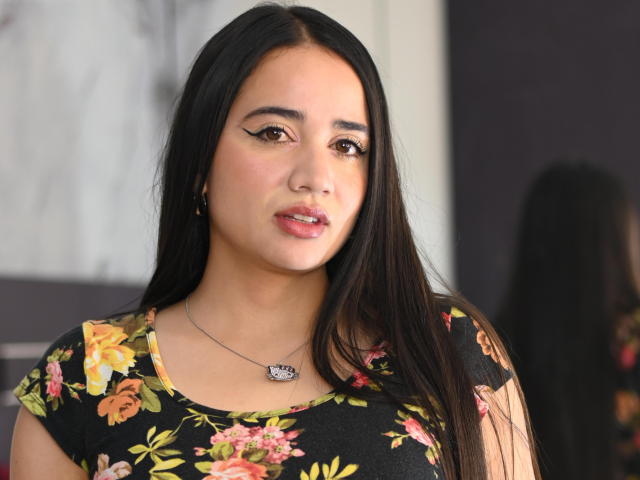 NicoleHolmes with long black hair, brown eyes, floral black top, pink lipstick, subtle eye makeup, necklace with purple gem, soft smile, slightly parted pink lips, light skin, looking forward.