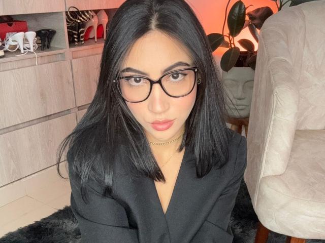 JuanaFox with black hair, glasses, and a black blazer, sitting on a furry black rug, has a gold necklace and a cute, serious expression.