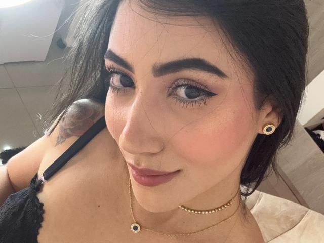 JuanaFox has dark hair, black eyeliner, and a subtle smile. She's wearing a black lace bra, gold necklace, and earrings. Tattoo on her right shoulder. Boobs visible.