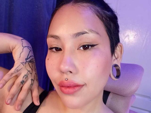 RebbekaBhons, con ojos marrones, cabello negro recogido en un moño, piercing en la nariz, delineador de ojos negro, labios rosados, aretes grandes, tatuajes en la mano izquierda, sentada en una silla..