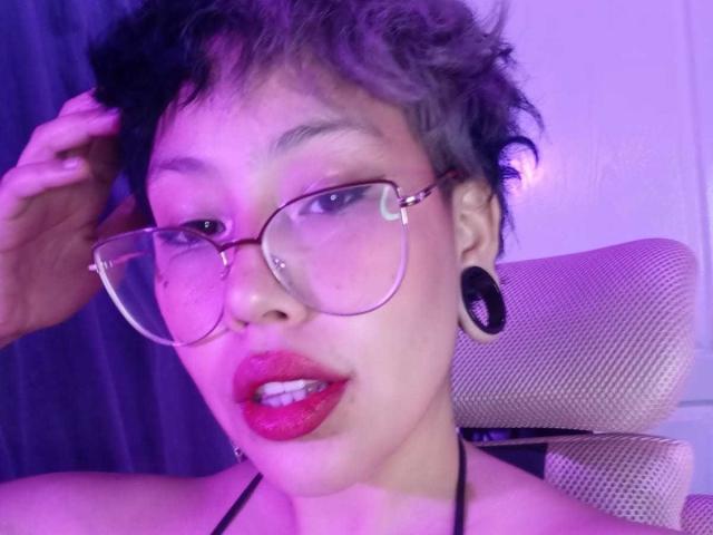 RebbekaBhons has short, curly grayish hair, wearing red lipstick, glasses, and black ear gauges, with a nose piercing, and a black halter top, looking slightly open-mouthed.