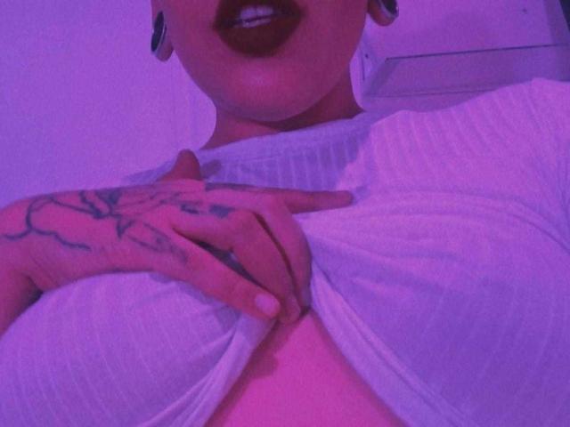 A mão de RebbekaBhons, coberta de tatuagens, segurava sua camisa branca listrada, puxando-a para baixo para expor seus seios grandes e rosados, usando batom escuro e alargadores de orelha pretos..