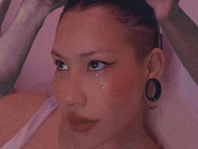 RebbekaBhons con un piercing en la nariz, delineador de ojos negro, una lágrima en la mejilla, cabello rojo recogido, aretes grandes negros, vistiendo una blusa rosa, labios ligeramente fruncidos..