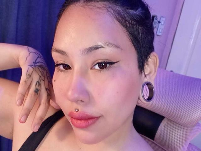 RebbekaBhons, con cabello oscuro recogido en un moño, delineador de ojos negro, piercing en la nariz, aro en el labio, camiseta negra, aretes grandes, tatuajes en la mano izquierda, sentada en una silla rosa..