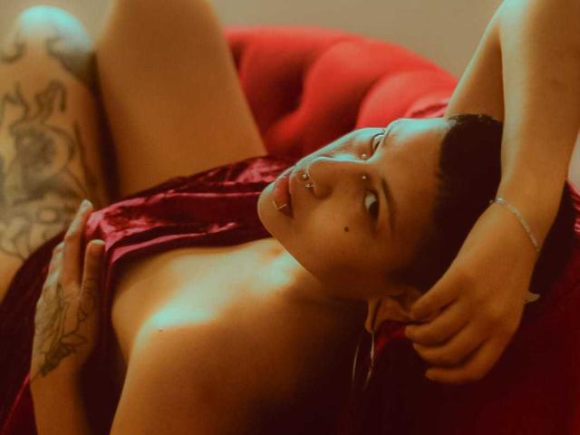 RebbekaBhons laying on a red chair, her hands on her chest, exposing her small tits, tattooed thigh, and piercing nose, with a seductive look and dark hair pulled back.