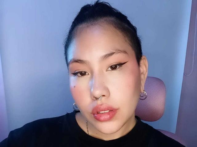 RebbekaBhons heeft een schattig gezicht met roze lippenstift, zwarte eyeliner, een neusring en oorringen. Ze draagt een zwarte top en zit in een roze stoel..