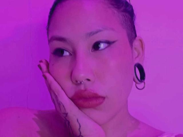 RebbekaBhons usa um piercing no nariz, alargadores pretos nas orelhas e tem tatuagens na mão, com a mão encostada na bochecha, em uma foto com iluminação roxa. Seus lábios estão pintados de vermelho..