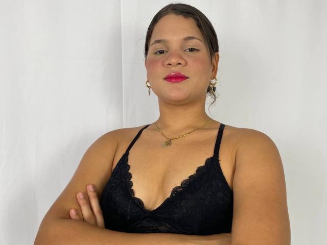 LatinTrioSex69, con los brazos cruzados, vistiendo un sujetador de encaje negro, lápiz labial rojo, aretes dorados y un collar, tiene senos medianos y una sonrisa segura..