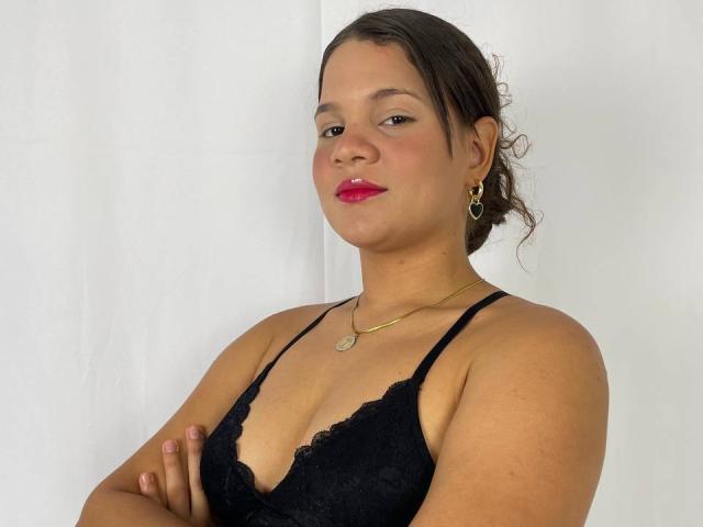 LatinTrioSex69, brown-haired, medium skin, red lipstick, black lace bra, gold necklace, earrings, arms crossed, slight smile, white background.