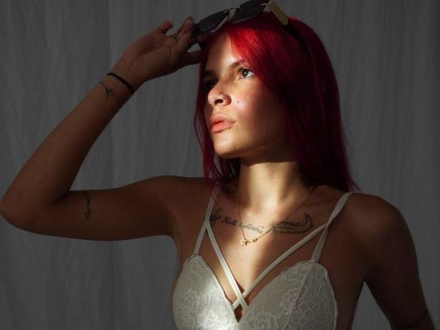 LatinTrioSex69 con cabello rojo, levantando gafas de sol, usando un sostén de encaje blanco, tatuajes en el brazo y el pecho, luz solar en la cara, mirando hacia arriba. Pechos visibles, escote sutil, expresión tranquila..