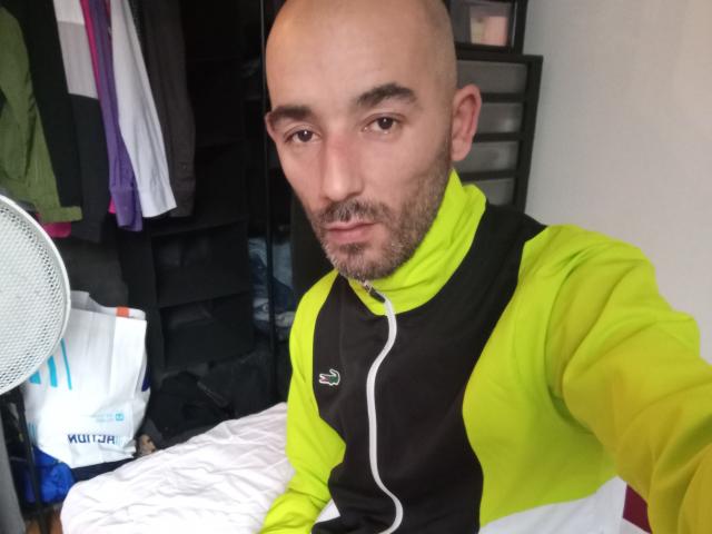 TeddyLeCoursier has a bald head, light brown beard, and is wearing a neon yellow and black sports jacket. He's sitting on a bed with a white sheet.