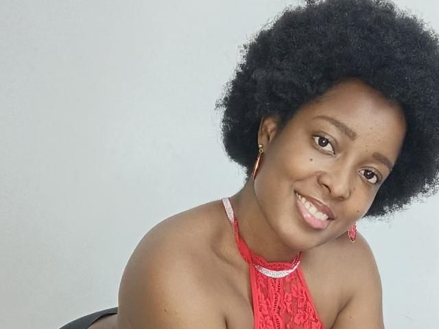 ChelseyeBonny with dark skin, natural afro, red lace halter top, red earrings, smiling at camera, showing small nose mole, upper body shot.