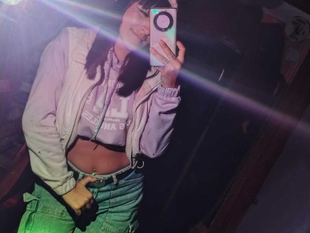 SydneySweetX in un selfie allo specchio, indossa una giacca bianca, pantaloni verdi e un top corto viola, con un reggiseno nero visibile, e tiene in mano un telefono con un anello..