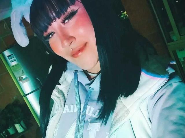 SydneySweetX met zwart haar, een pony en witte konijnenoren, draagt een licht jasje, heeft roze blush, een cat-eye eyeliner, een neusring en een schattende glimlach, en laat haar borsten zien..