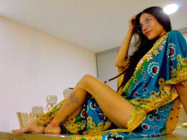MiaUtena sits on a glass table, wearing a blue and yellow patterned sari, showing her bare ass, long black hair, glasses, and a tattoo on her left thigh.