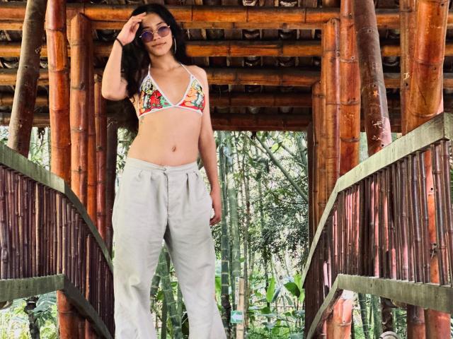 MiaUtena with long dark hair, wearing a colorful bikini top and white pants, standing in a bamboo structure, touching her sunglasses, showing a flat stomach and small tits.