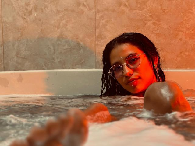 MiaUtena with wet black hair, wearing glasses, in a bathtub, her brown tits and arm visible, red-orange light, water, and marble tiles in the background.