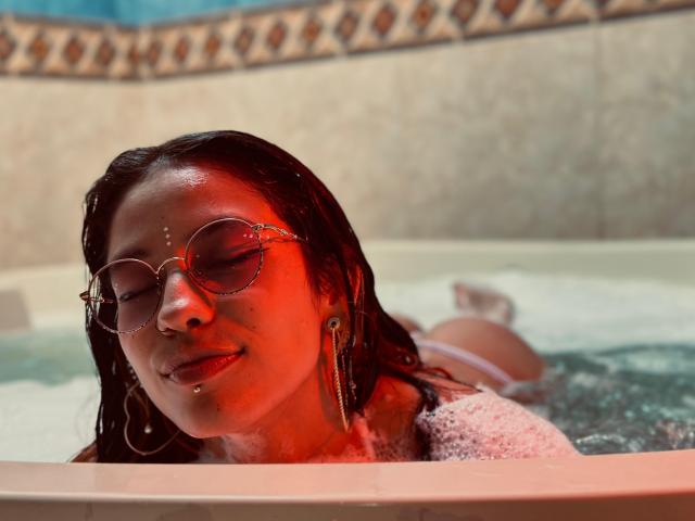 MiaUtena with wet brown hair, red light on face, wearing round glasses and gold earrings, lying in a bubbly bathtub, eyes closed, showing a nose ring and a lip piercing.