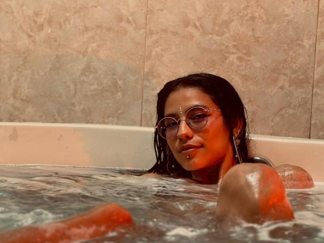 MiaUtena in a bathtub, water up to her shoulders, wearing round glasses, with wet black hair, looking at the camera, her bare boobs partially submerged, marble tile background.