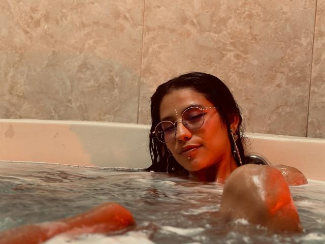 MiaUtena with wet black hair, wearing glasses and a nose ring, has her eyes closed in a bathtub, her shoulder and arm visible above the water.