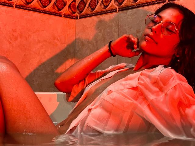 MiaUtena in a red-lit bath, wearing a wet, open shirt, with glasses, dark hair, and a black bracelet, her hand near her mouth, in water. Her tits are visible.