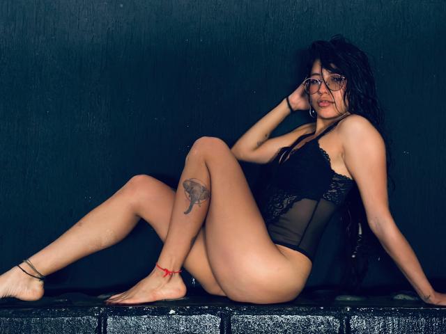 MiaUtena with wet black hair, glasses, and a lace black bodysuit, sitting on black bricks, showing a tattoo on her right thigh and a red anklet.