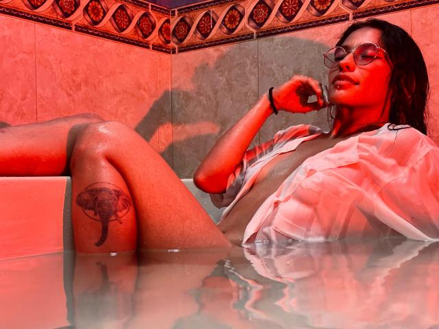 MiaUtena with wet shirt, red lighting, glasses, and tattoo on thigh, lounging in a tub, legs up, hand near mouth, water reflecting her. Red shadows on tan tiles.