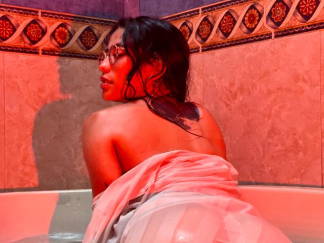 MiaUtena, naked, in a bathtub with wet, pinkish skin, back to camera, glassy eyes, dark wet hair, white towel around ass, red lighting, tattoos on upper back.