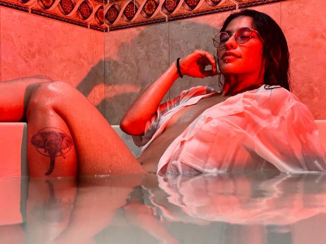 MiaUtena with wet skin, red lighting, glasses, and an open shirt, sitting in a bathtub, showing a tattoo on her thigh and a slightly visible pussy.