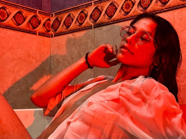 MiaUtena with red-tinted skin, wearing glasses, white shirt, and black bracelet, holding phone to ear, sitting with legs up, in a bathroom with decorative tile.