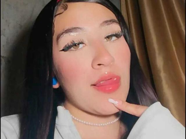 SydneySweetX has glossy pink lips, long black hair, thick black eyeliner, and a pearl necklace. She's touching her chin with white-manicured fingers, looking at the camera.