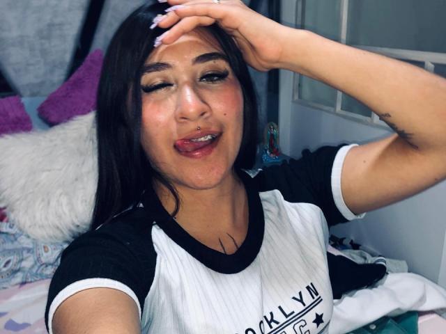 SydneySweetX with dark hair, winking, tongue out, black and white shirt, hand on head, nose ring, tattoo on arm, and a ring on her finger. Bedroom setting.