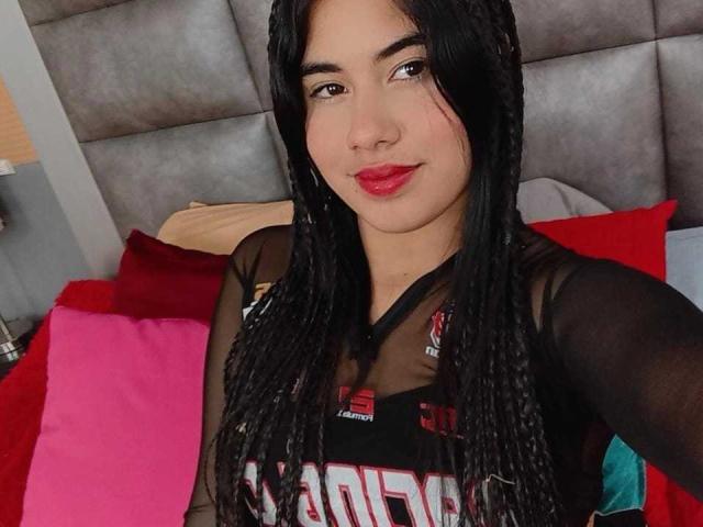 BiancaSolas has long black braids, red lipstick, and a black sheer top showing her small tits. She's smiling with red and pink pillows behind her.