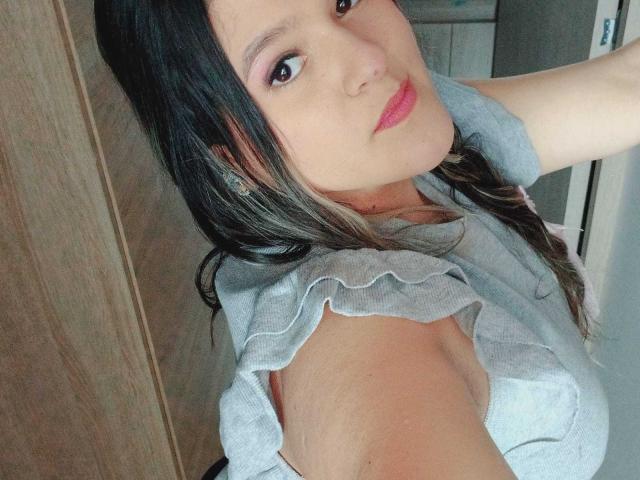 MiaJhonss with light pink lipstick, gray ruffled top, showing her tits, brown hair, and a slight smile, holding arm up, wearing small diamond earrings.