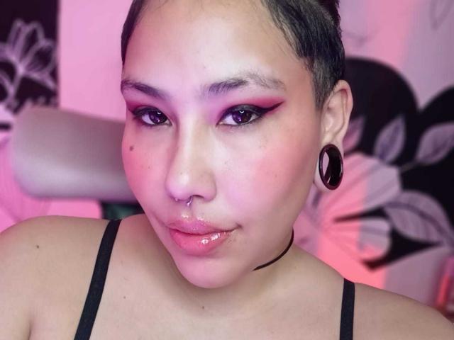 RebbekaBhons tiene el cabello corto y negro, sombra de ojos rosa, lápiz labial morado, labios rosados brillantes, aretes negros, collar negro, piercing en la nariz y una camiseta negra, con iluminación rosa y morada..
