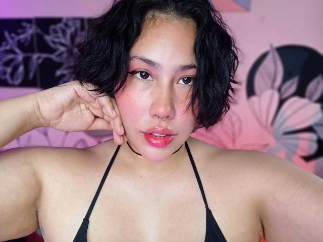 RebbekaBhons with short black hair, pink eyeshadow, nose ring, and glossy pink lips, wearing a black halter top, showing her tits. She's touching her cheek with one hand.