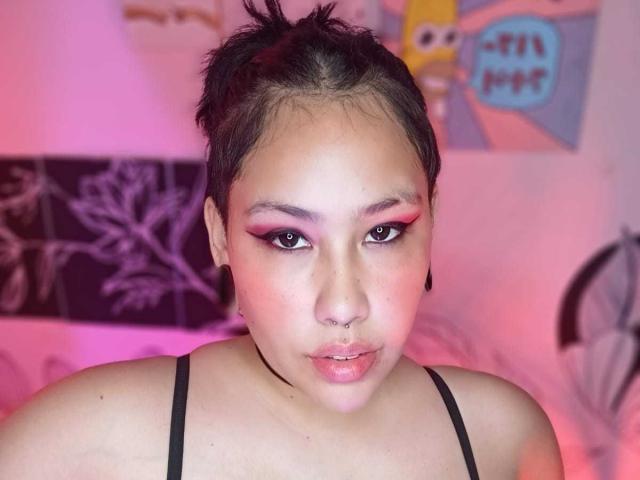 RebbekaBhons, con cabello corto y negro, sombra de ojos rosa, un sostén negro, un piercing en la nariz y expansores negros en las orejas, mirando a la cámara con un ligero puchero. Iluminación rosa y morada..
