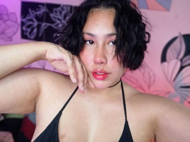 RebbekaBhons has curly black hair, brown eyes, nose ring, and red lips. She's wearing a black halter top, showing her small tits, with a tattoo on her right hand.